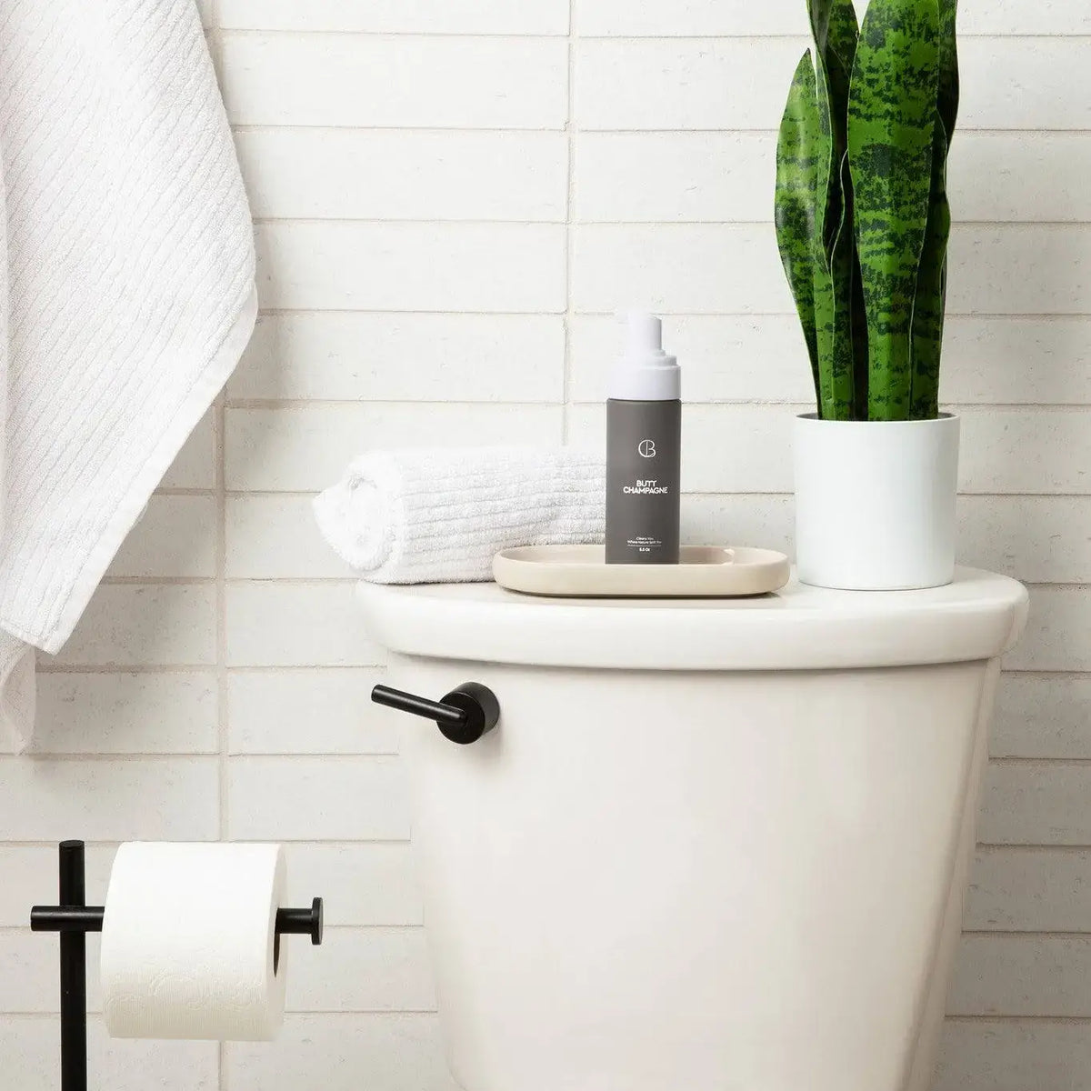 Bottle of soap on a toilet tank with a plant and towel in a bathroom setting