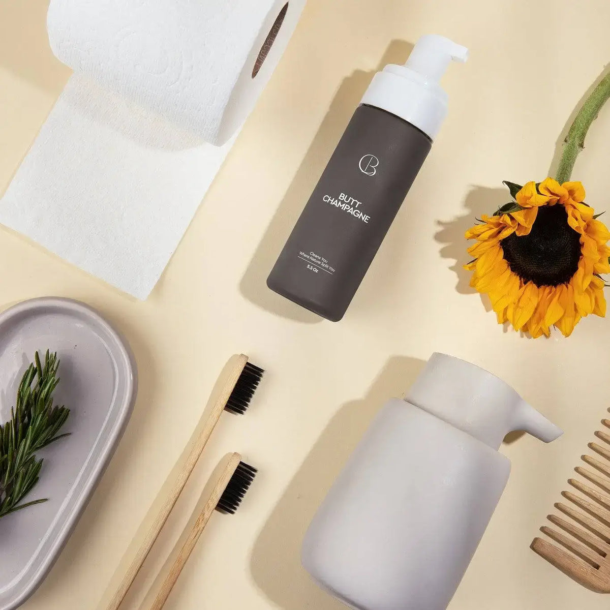 Bottle of Butty Champagne with a sunflower, bamboo toothbrush, and ceramic container on a beige surface.