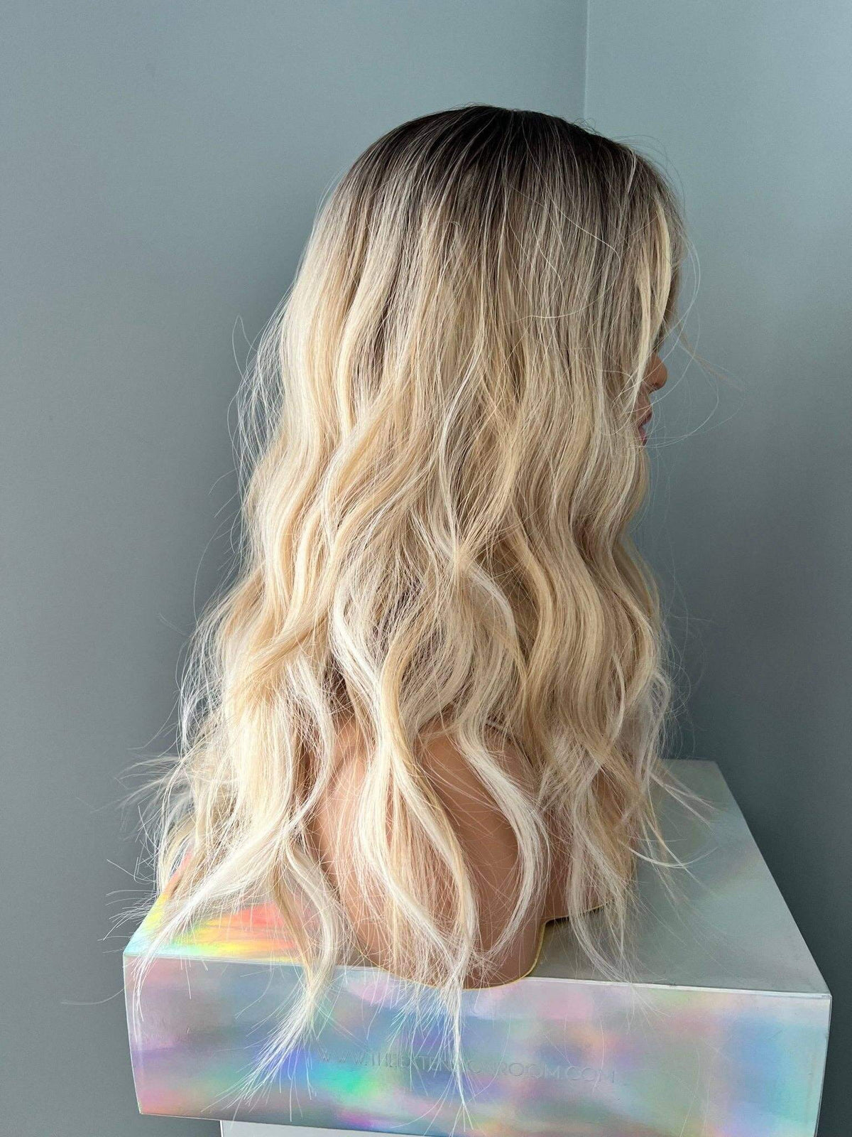 Person with long, wavy blonde hair standing against a plain wall.