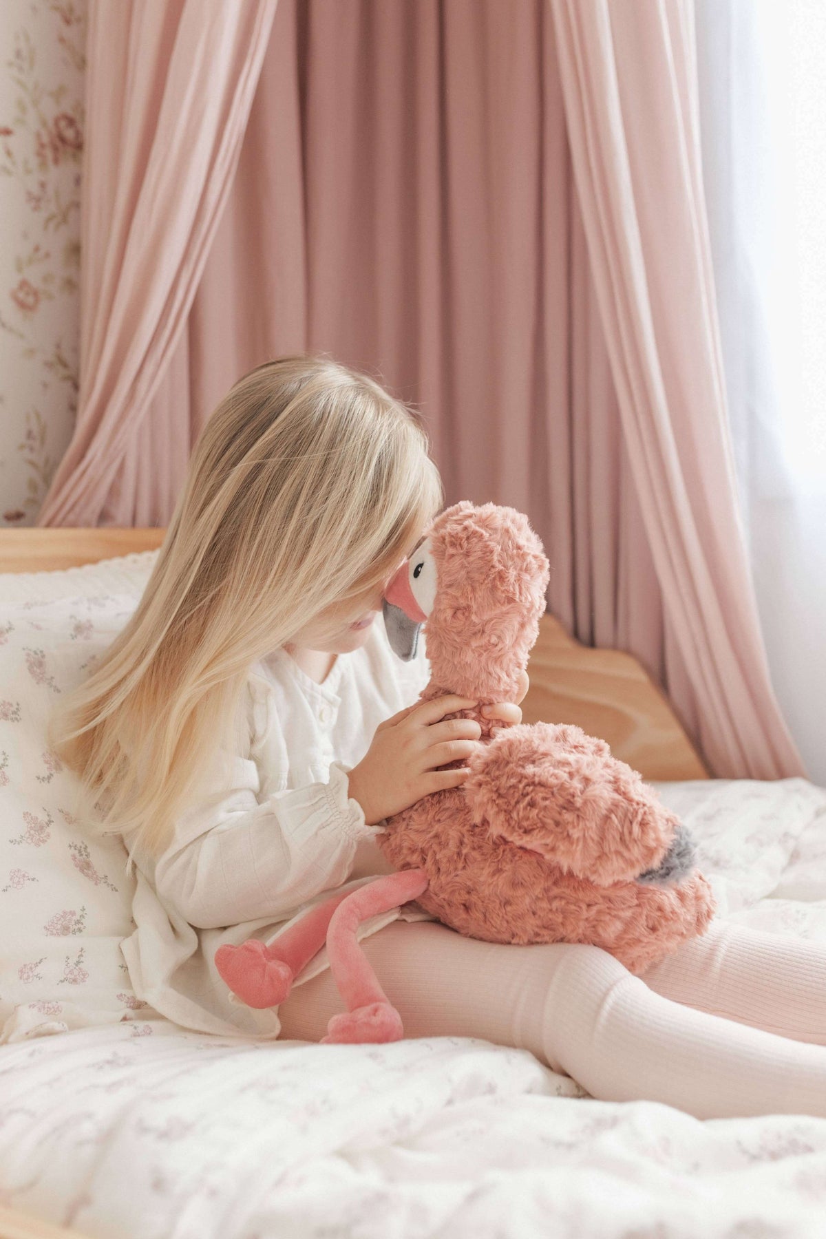 A child cuddling Francesca The Flamingo Weighted Teddy plush toy on a cozy bed.