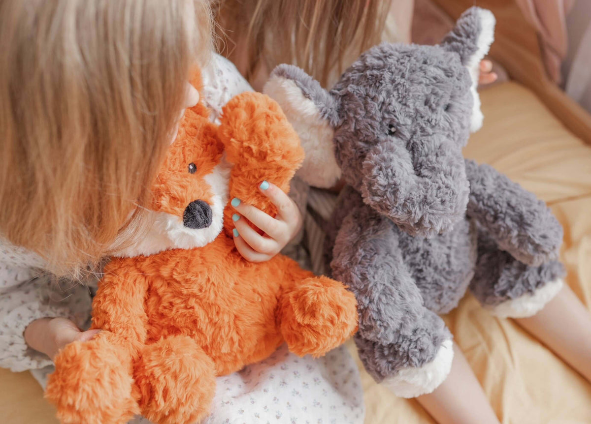 Children holding Frankl The Weighted Fox teddy and a plush elephant toy on a cozy bed.