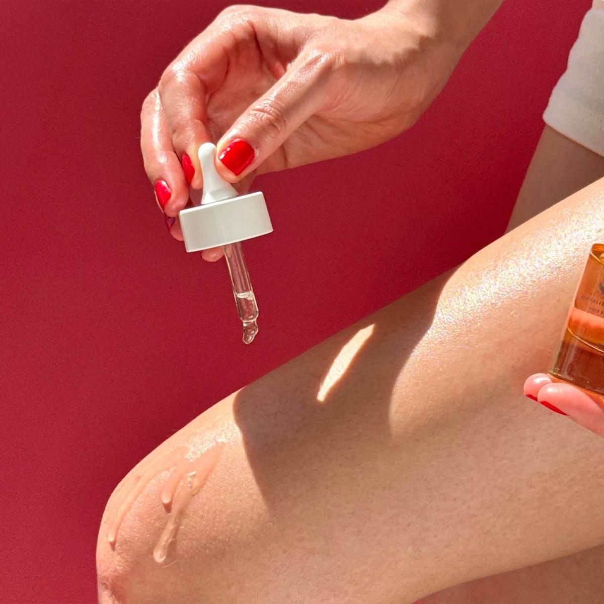 Hand applying a dropper of skincare product to skin against a red background