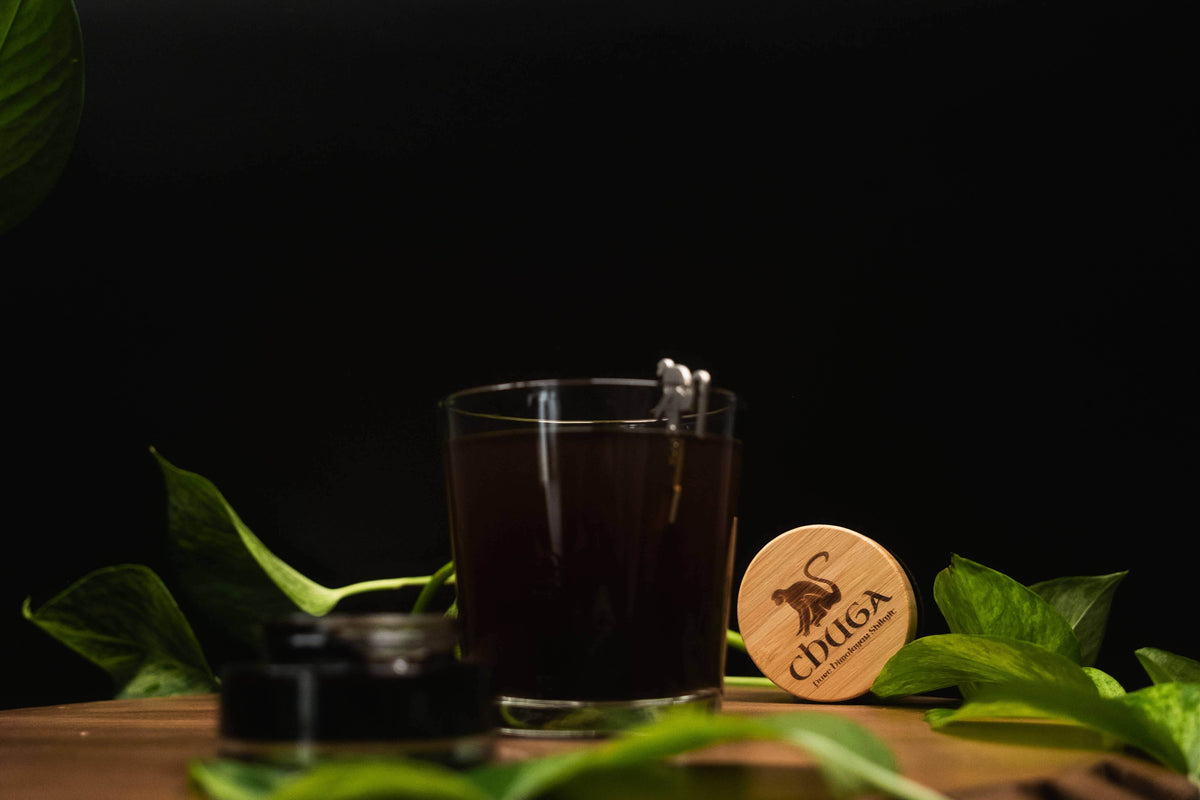 Glass of dark liquid with a wooden coaster featuring 'Iduga' brand on a dark background with green leaves.