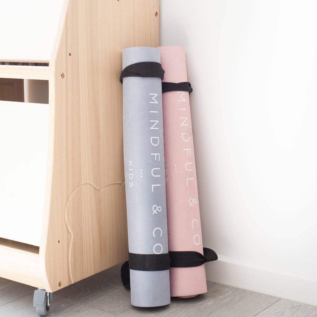 Two yoga mats, one gray and one pink, with 'Mindful & Co' branding, leaning against a wooden piece of furniture.