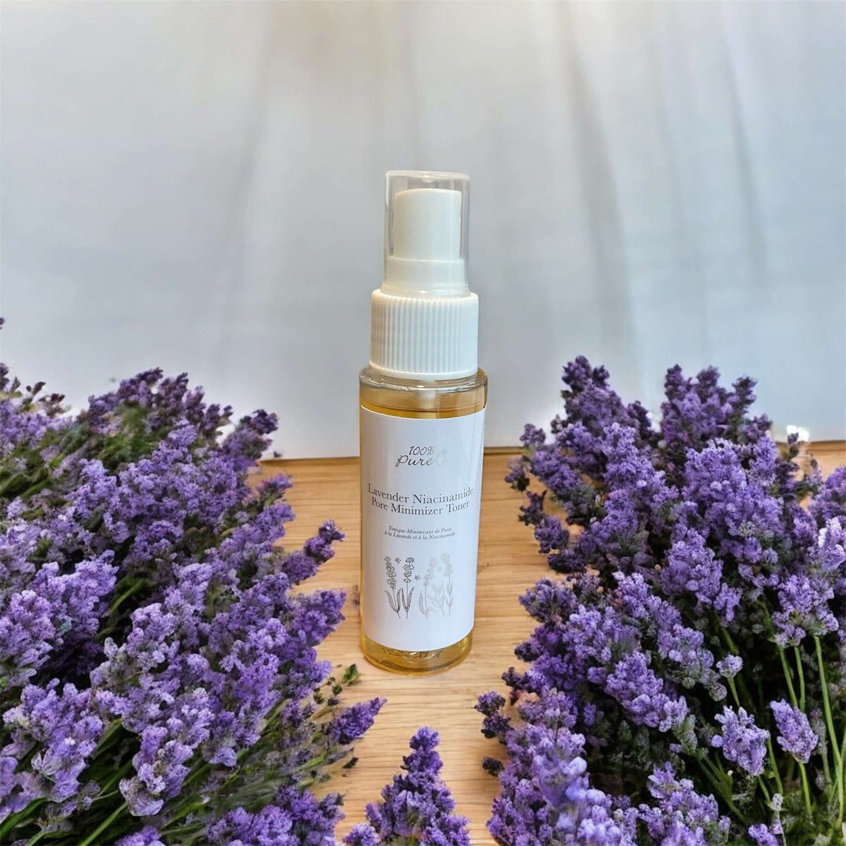 Spray bottle with lavender essential oil surrounded by lavender flowers on a wooden surface.