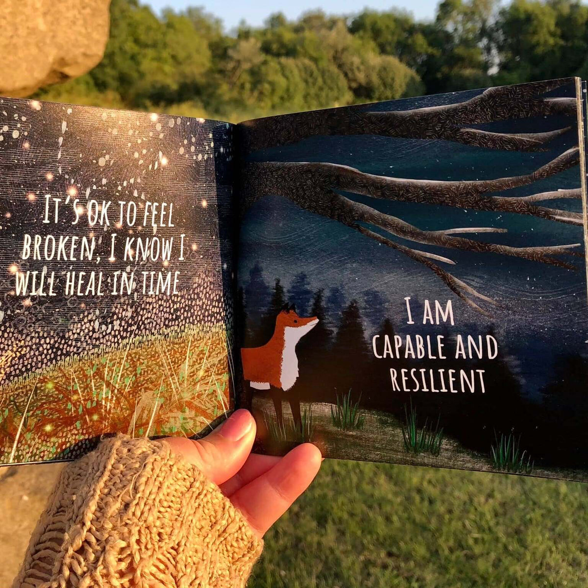 Open book with motivational text and illustrations held by a person outdoors.