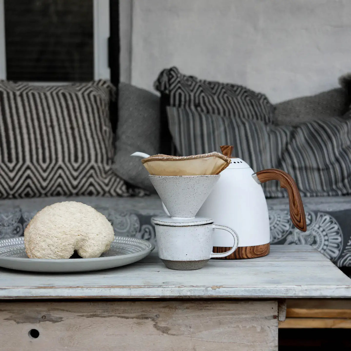 White ceramic coffee maker with wooden handle on a wooden table, with a sofa and cushions in the background.
