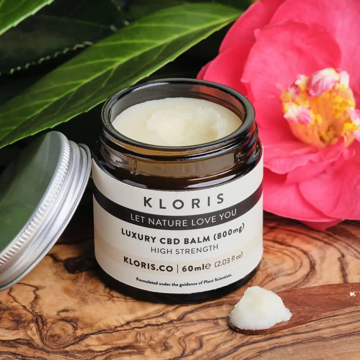 Kloris CBD balm jar on a wooden surface with a pink flower and green leaves in the background