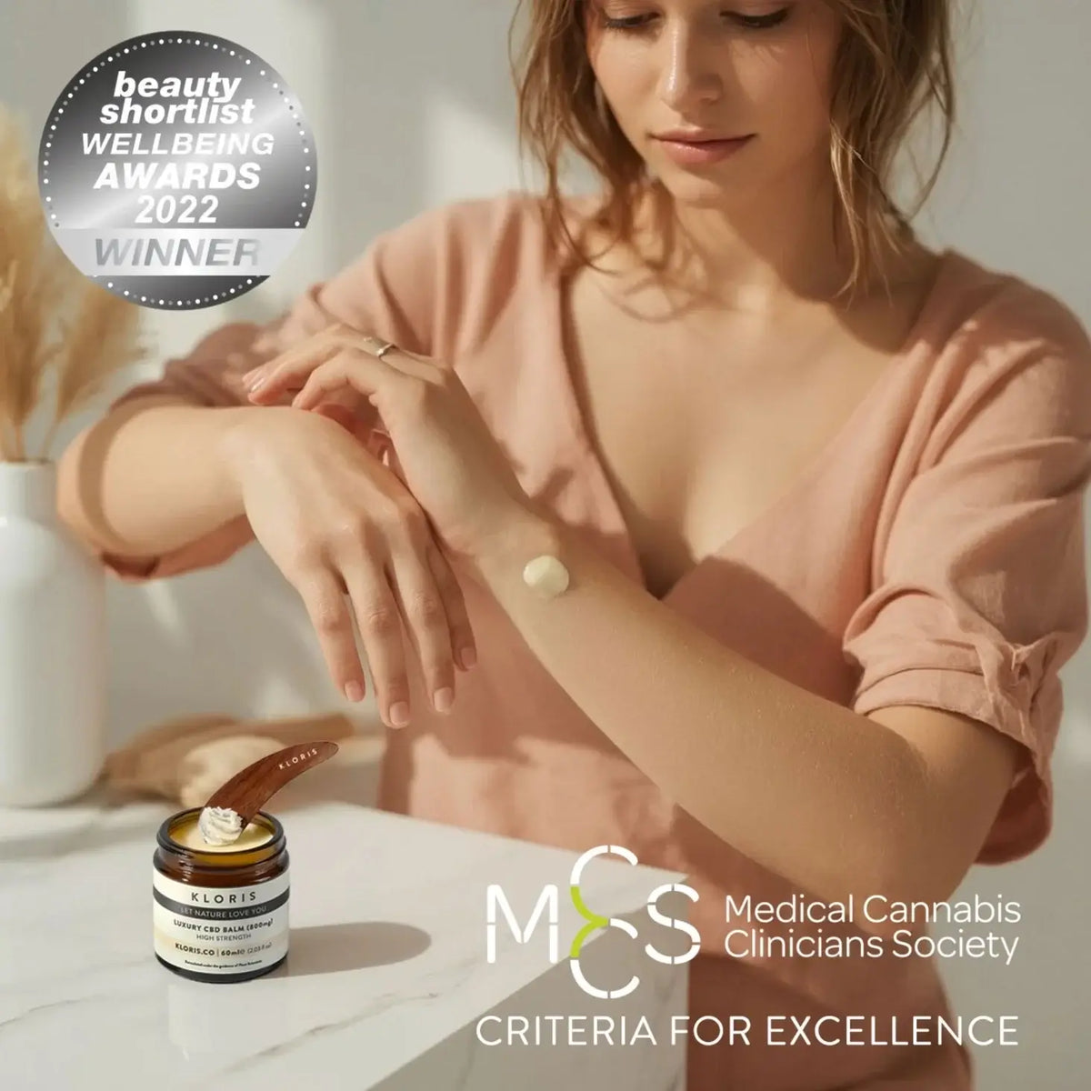 Woman applying cream to her wrist with a jar of cream on a table, surrounded by 'Medical Cannabis Clinicians Society' and 'Beauty Shortlist Wellbeing Awards 2022' text.