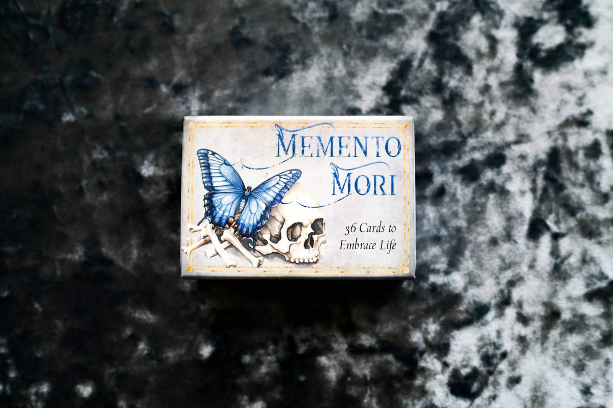 Memento Mori Cards packaging featuring a blue butterfly and skull design on a textured background.
