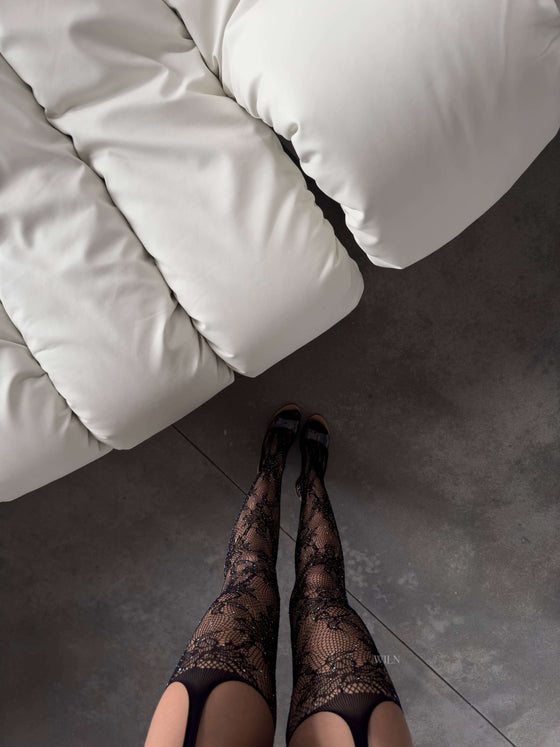 Person wearing black lace stockings standing on a gray floor next to a white couch.