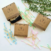Cotton buds with colored tips in cardboard boxes on a white surface with greenery.