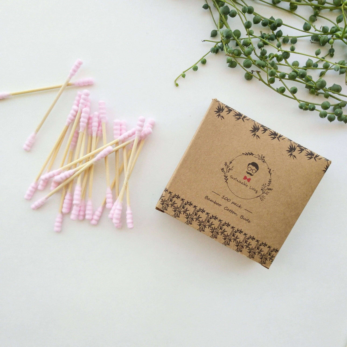 Box of cotton buds with pink tips on a white surface with greenery