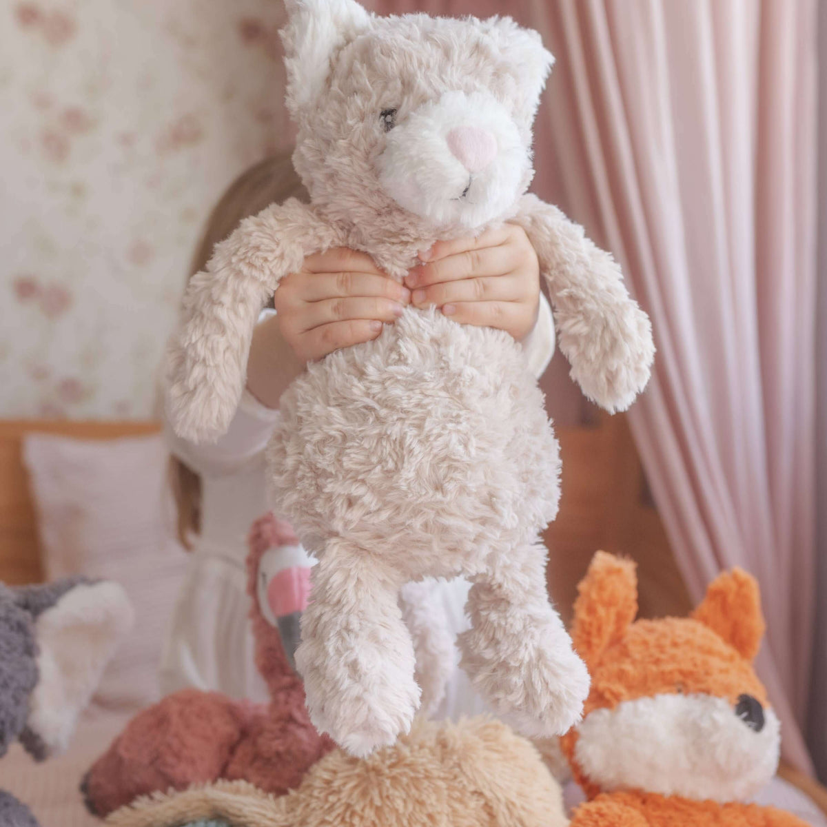 Stack of plush toys including a cat, bear, and fox on a bed with a soft pink curtain in the background.