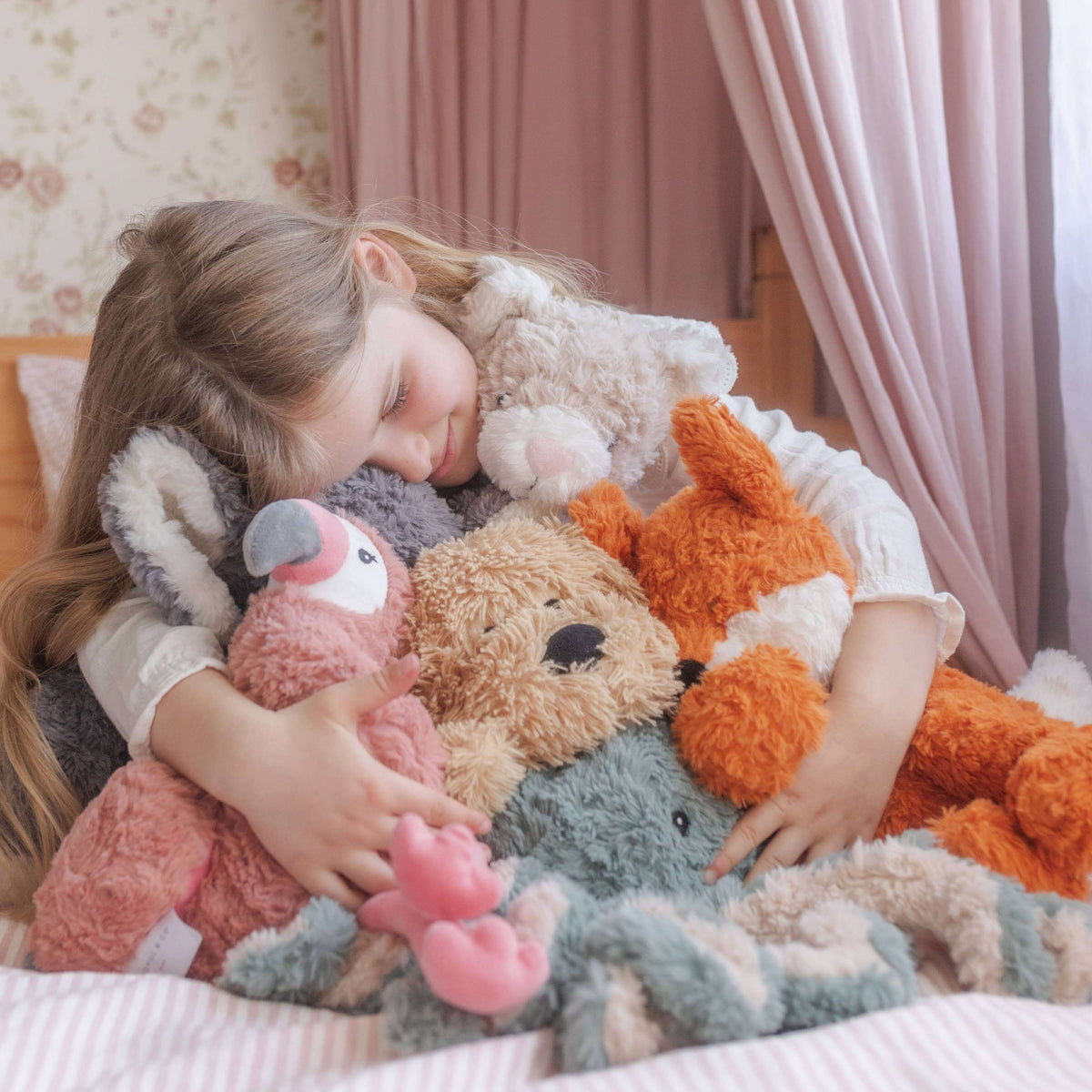 Child hugging a teddy bear and other plush toys in a cozy room.