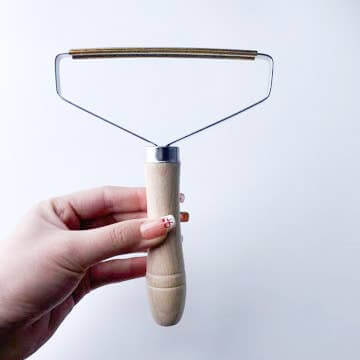 Hand holding a Plastic-Free Travel Lint Remover with a natural wooden handle and durable metal edge.