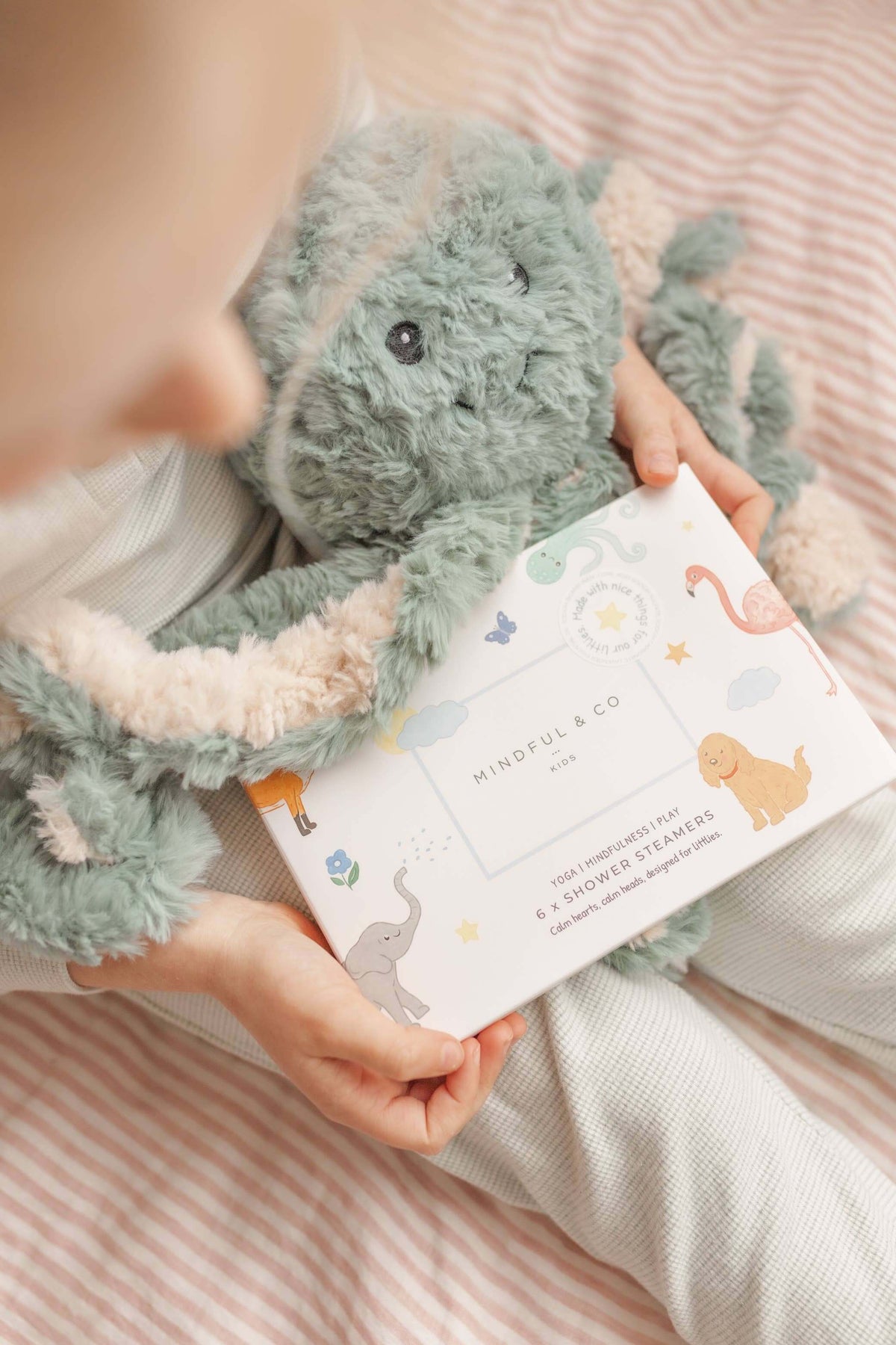 Child holding a pack of Sensory Shower Steamers x 6 next to a plush toy on a striped blanket.
