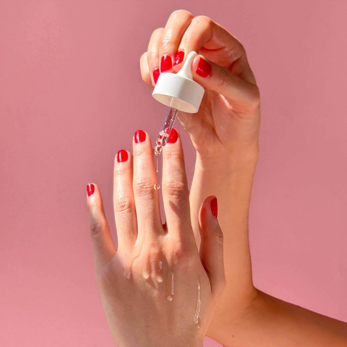 Hand applying clear nail polish from a bottle with a pink background