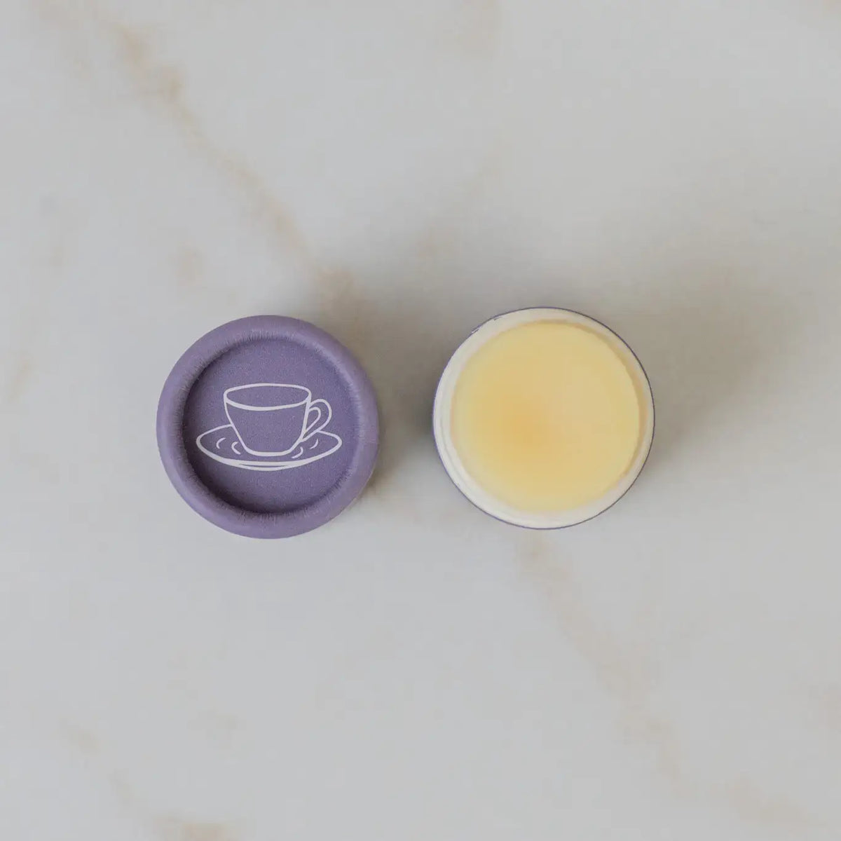 Round container with purple lid featuring a tea cup design, filled with a yellowish substance on a light marble background.