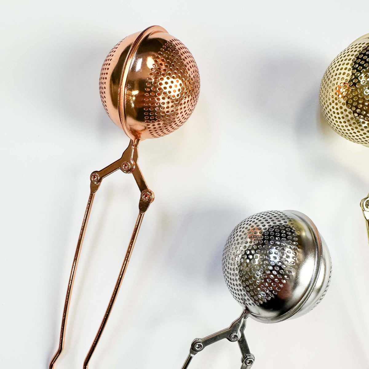 Metallic tea infusers in rose gold, silver, and gold on a light background