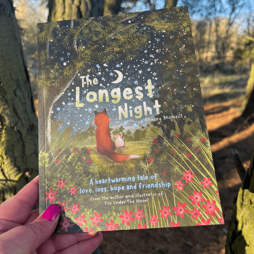 The Longest Night Paperback Book + bookmark held in hand with forest background, showcasing the cover design.