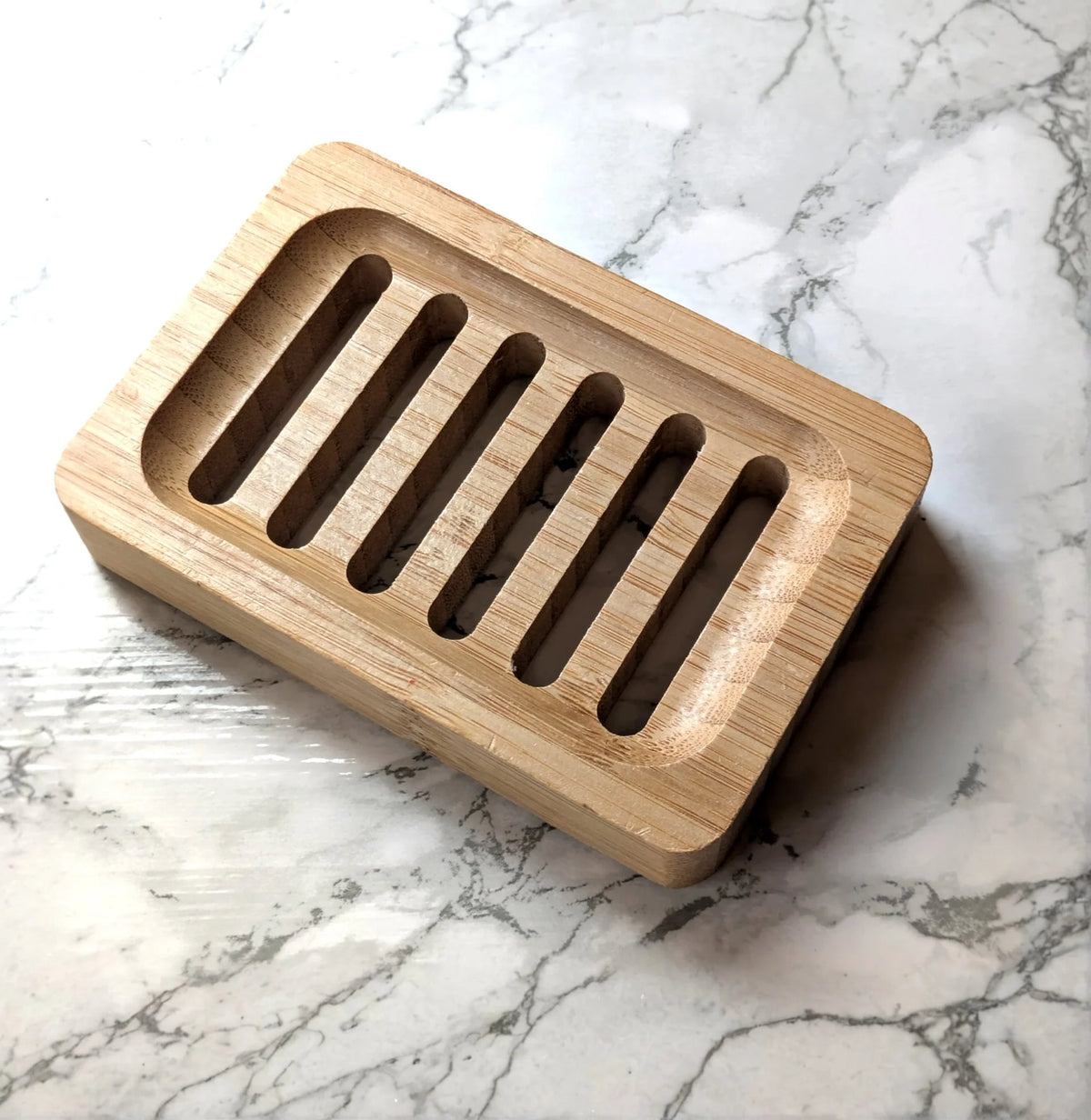 A rectangular bamboo soap dish with five elongated cutouts, photographed on a white marble surface with gray veining.