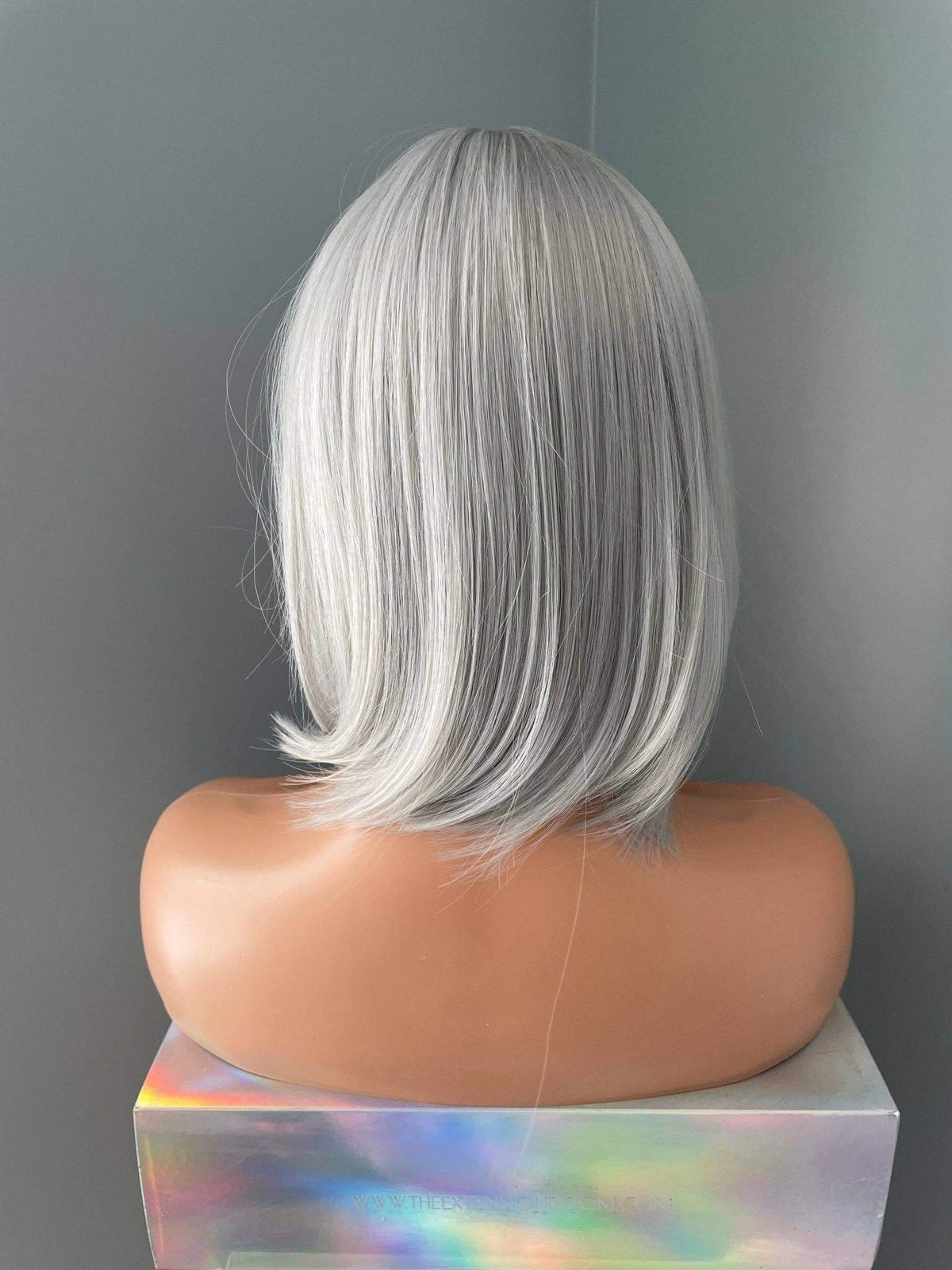 A short, straight wig with silver-gray and white highlights, shown from the back on a mannequin head mounted on a holographic display block against a gray background.