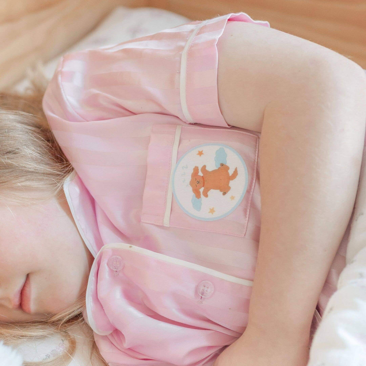 A close-up of a child wearing a shiny light pink pajama top with a decorative chest patch showing a cartoon dog, lying down in a soft bed setting.